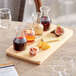 An Acopa natural wood flight tray with glass carafes of brown liquid on a table with wine and cheese.