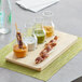 An Acopa natural wood flight tray with skewers of meat and sauces on it.