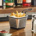 A small black wire basket with a handle, lined with white paper and filled with French fries, sits on a wooden counter.