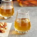 Two Acopa whiskey glasses filled with amber liquid on a table with a plate of nuts.
