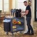 A man standing next to a Cambro charcoal gray service cart with food on it.