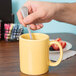 A hand holding a spoon in a yellow Tuxton C-handle mug.