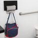 A blue and red bag hanging from a Bobrick stainless steel mobile device holder on a white wall.
