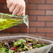 A hand pouring yellow liquid into a bowl of salad using a Tablecraft Marbella oil & vinegar cruet.
