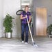 A woman in a purple shirt using a ProTeam ProBlade carpet floor vacuum tool attached to a backpack vacuum cleaner.