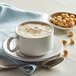 A white cup of UPOURIA hazelnut cappuccino with foam and nuts on the side.