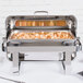A Vollrath stainless steel roll top chafer on a large silver tray with food inside.