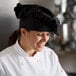 A woman wearing a black Mercer Culinary chef's hat smiles while cooking at a professional kitchen counter.