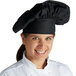 A woman wearing a Mercer Culinary black soft chef hat while smiling in a professional kitchen.