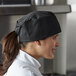 A woman in a Mercer Culinary black Baker's skull cap smiling in a professional kitchen.