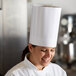 A woman in a Mercer Culinary white chef's hat smiles while holding a knife.