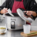 A person in a chef's uniform holding a white non-stick pot lid over an Avantco rice cooker.