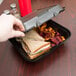 A hand taking out a sandwich from a Genpak black foam container.