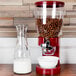 A red Zevro dry food dispenser filled with cereal next to a white bowl of cereal and a glass of milk.