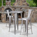A Flash Furniture silver metal bar height table and two chairs on an outdoor patio in front of a stone fireplace.