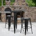 A Flash Furniture black metal bar height table with two square black stools on a stone patio.