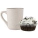 A white frosted cupcake in a white fluted baking cup next to a mug.