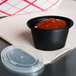 A Newspring black oval souffle container with red sauce and a clear lid.