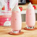 Two glasses of pink strawberry milkshakes on a wooden table with a bag of Bossen Strawberry Powder Mix.