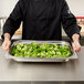A person in a black coat holding a Vollrath Miramar decorative rectangular food pan filled with broccoli.