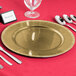 A round gold metal charger plate with a beaded rim on a table with silverware and a glass.