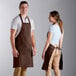 A man and woman standing next to each other at a counter in a professional kitchen, both wearing brown Choice bib aprons.