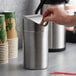 A hand putting a paper into a Vollrath stainless steel mini waste can on a counter.