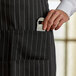 A person's hand holding a bottle opener in a pocket of a black and white pinstripe Tuxedo Apron.