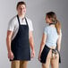 A man and woman standing in a professional kitchen. The man is wearing a black apron and the woman is wearing a blue apron.