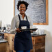 A smiling woman wearing a navy blue Choice bib apron holding a tray of coffee cups.