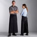 A man and woman standing on either side of a counter wearing black Choice full-length bistro aprons.