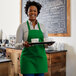 A smiling woman wearing a Choice Kelly Green poly-cotton bib apron holding a tray of coffee cups.