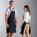A man and woman standing next to each other wearing navy blue bib aprons with pockets.