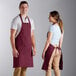 A man and woman wearing burgundy bib aprons standing next to each other at a counter.