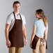 A man and woman standing on a counter in a professional kitchen wearing brown Choice poly-cotton bib aprons.