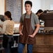 A man wearing a Choice brown bib apron with 3 pockets standing in a coffee shop.