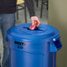 A man using a Rubbermaid BRUTE blue recycling bin lid with a mixed recycle slot to put a can in a blue trash can.