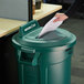 A hand putting paper into a green Rubbermaid recycling bin lid with a paper slot.