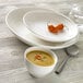 A bowl of soup and a spoon on a Libbey Royal Rideau white deep coupe plate.