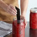 A hand using a black Ball canning utensil to cut tomatoes in a jar of red sauce.