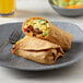 A plate with a breakfast burrito made with Mission 12" Chipotle Tortillas, vegetables, and cheese, with a bowl of fruit on the table.