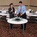 A man and woman standing in a room with a Lifetime white granite plastic folding table and chairs.