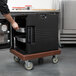 A person using a dark brown Cambro Camdolly to move a black food container.