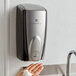 A close-up of a Rubbermaid black and grey automatic soap dispenser with white foam on a person's hand.