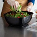 A woman wearing gloves preparing a salad in a black Fineline Super Bowl Plus container.