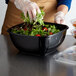 A woman in gloves preparing a salad in a Fineline black plastic bowl.
