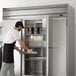 A man wearing a black apron and gloves opening a Beverage-Air Horizon Series reach-in refrigerator with stainless steel doors.