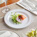 A Visions white plastic plate with a salad and fork on it on a table with a white cloth and other place settings.