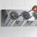 A hand holding a spoon in a Steril-Sil stainless steel flatware organizer with gray perforated cylinders.