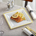 A Visions plastic square plate with gold bands holding a plate of food with a slice of lemon on top.